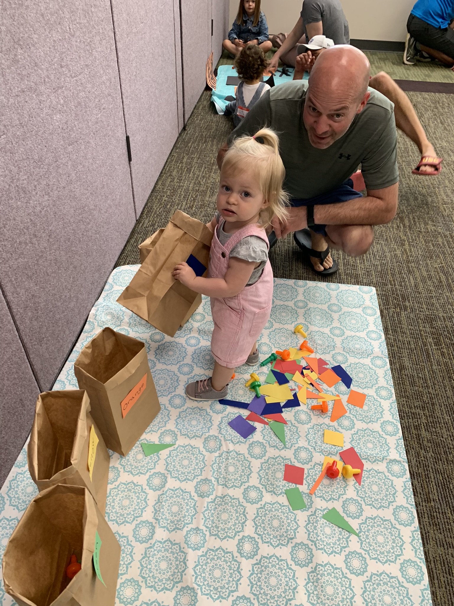 Toddler standing on table cloth sorting colored paper into paper lunch bags