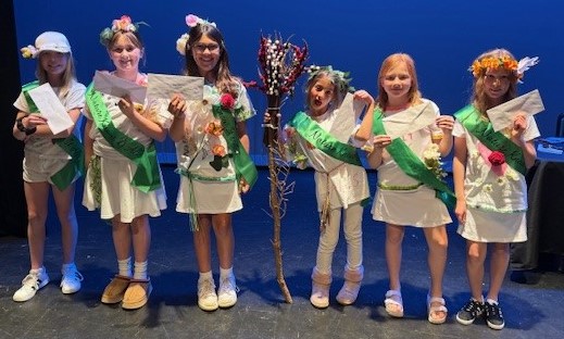 Six girls in white skirts and pants with green sashes and flowers on their shirts.