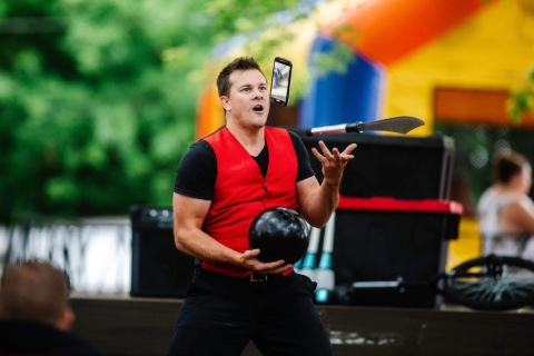 Man in red vest juggling a bowling ball, cell phone and bowling pin.