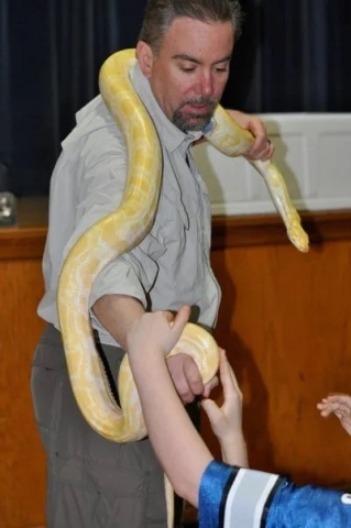 Man holding large yellow snake around his neck with a child touching it.