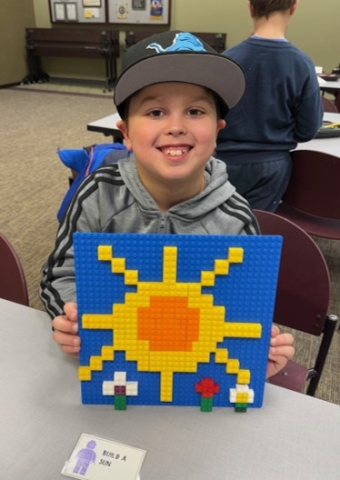 Boy with baseball cap smiling holding a Lego creation of a sun