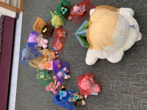 Stuffed animals with scarves on their heads. A big bear is reading a book to the other stuffed animals.