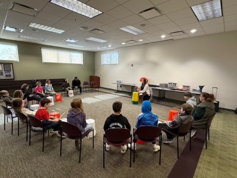 Group of older kids sitting in a semi-circle with large buckets on the floor in front of them and drumsticks in their hands. An instructor is up front.