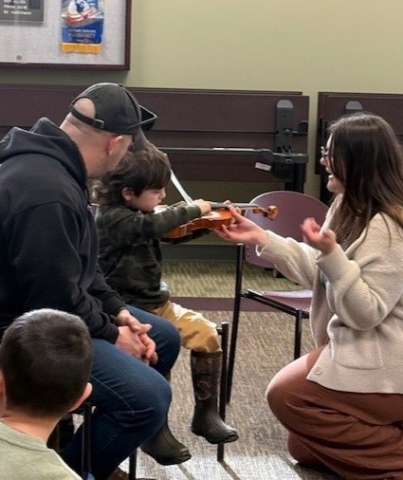 Preschooler playing violin on his dad's lap, with the instructor kneeling down helping.